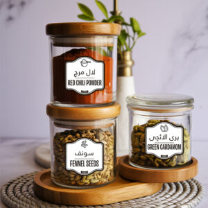 Glass jars with bamboo lids featuring pantry labels for spices – red chili powder, fennel seeds, and green cardamom – labeled in English and Urdu.